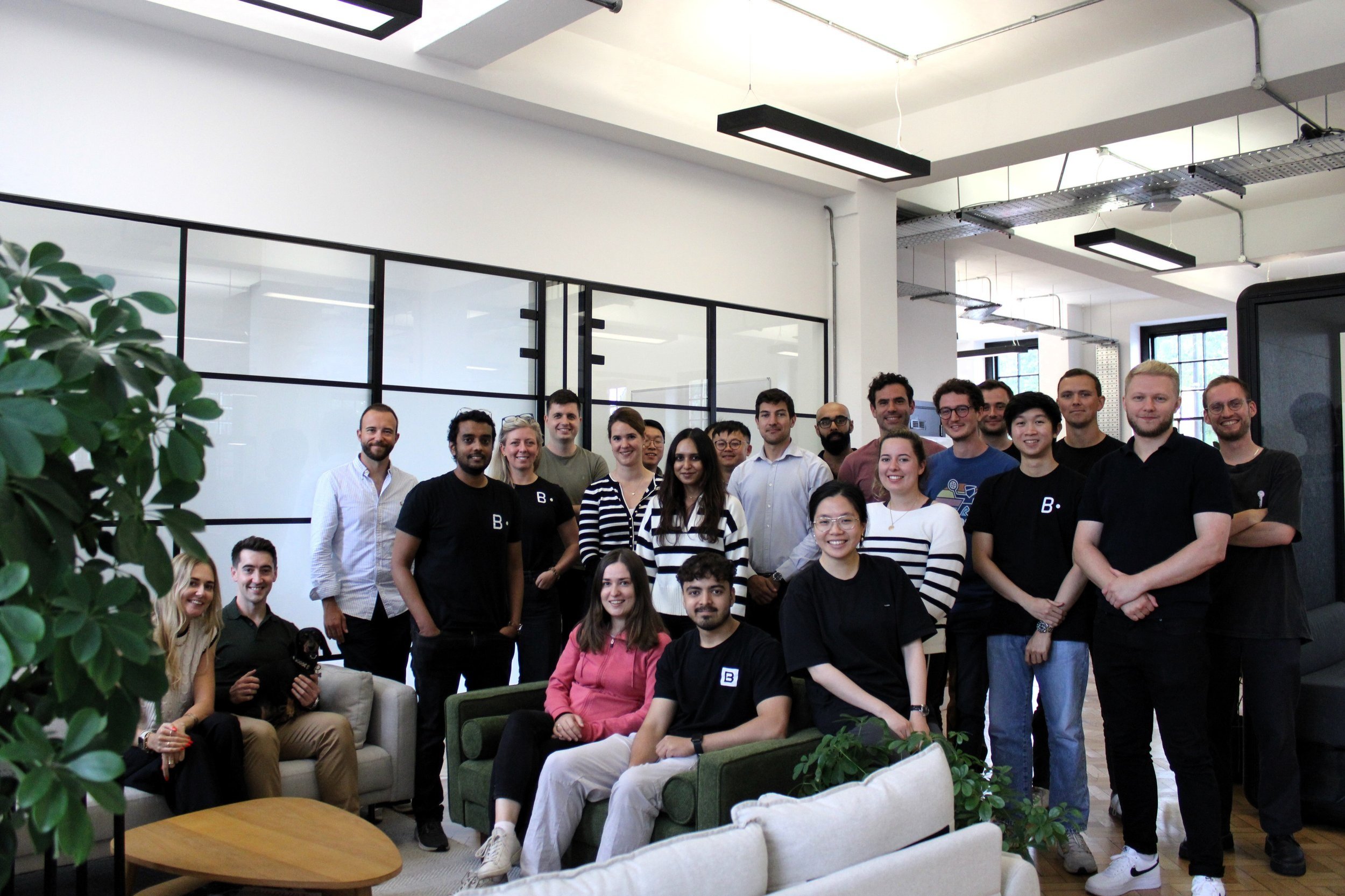 Group of diverse young professionals gathered in a modern office space with large windows and glass partitions.