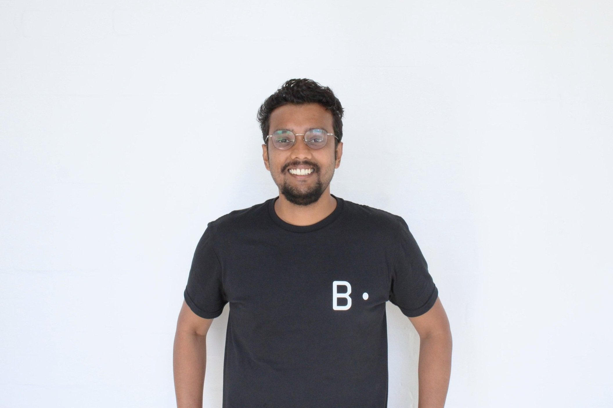 A man with glasses and a beard smiling, wearing a black t-shirt with a white letter B and dot on it, standing against a plain white wall.