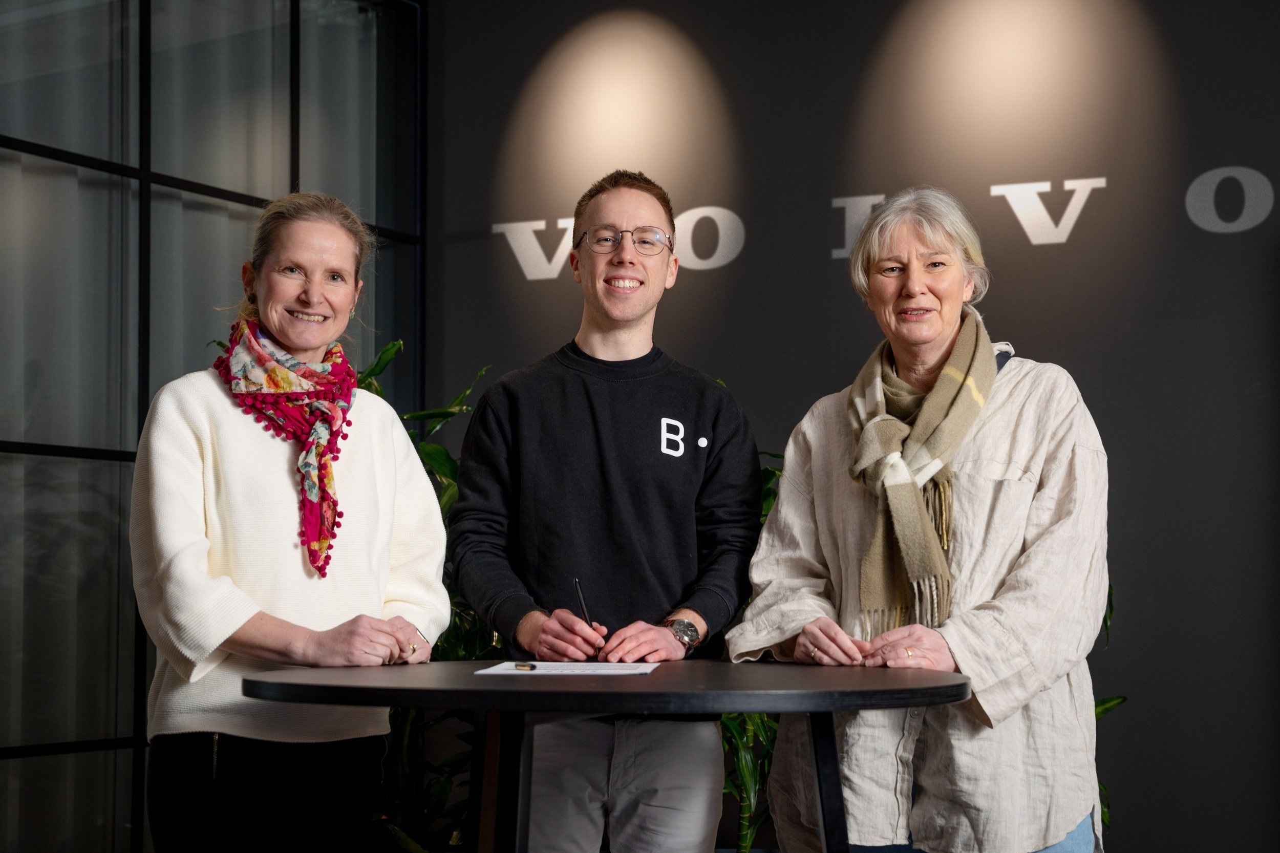 Three individuals standing behind a small table with a document and pen, smiling, in front of a dark wall with the word 'Vovo' in large white letters and greenery in the background.