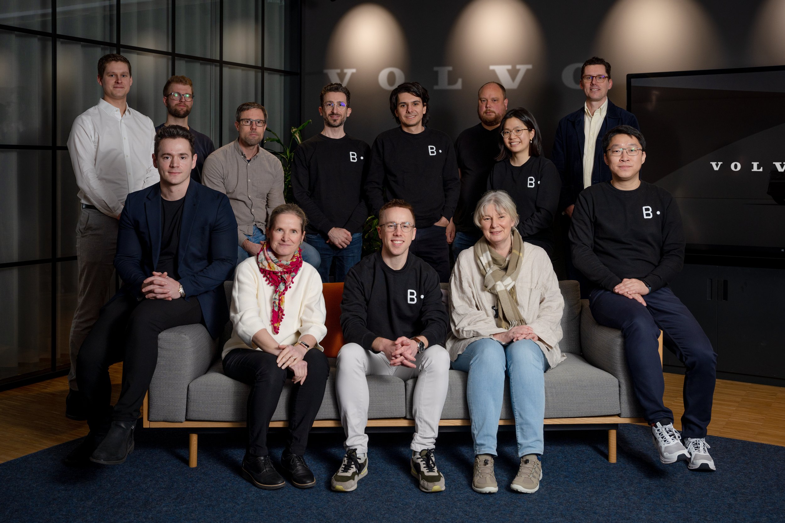 A group of fifteen people posing for a photo in a modern office space with the Volvo logo in the background. Some are seated on a gray sofa, and others are standing behind it, smiling.