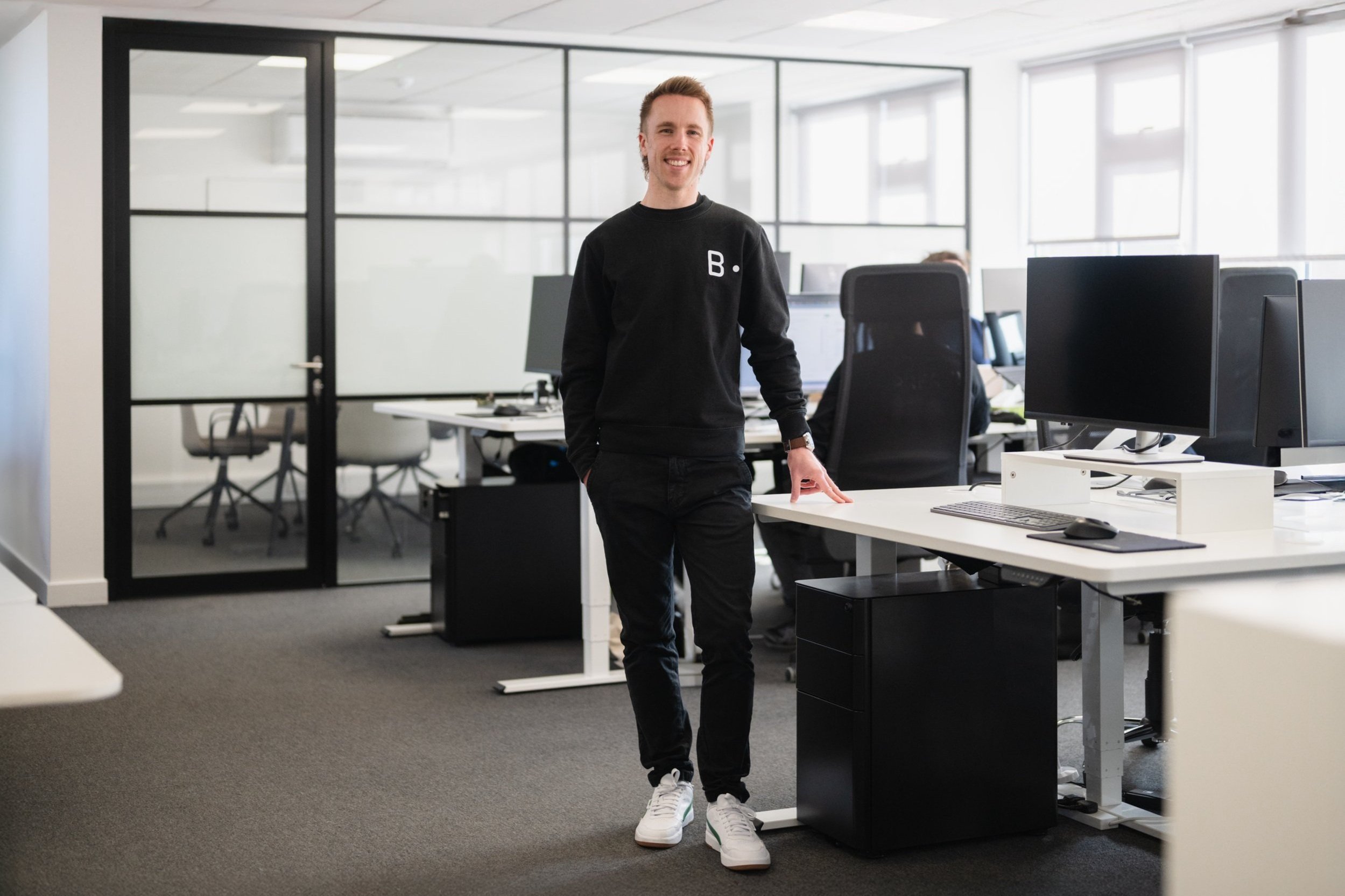 Young man standing in an open office workspace with computers and ergonomic chairs, smiling at the camera.