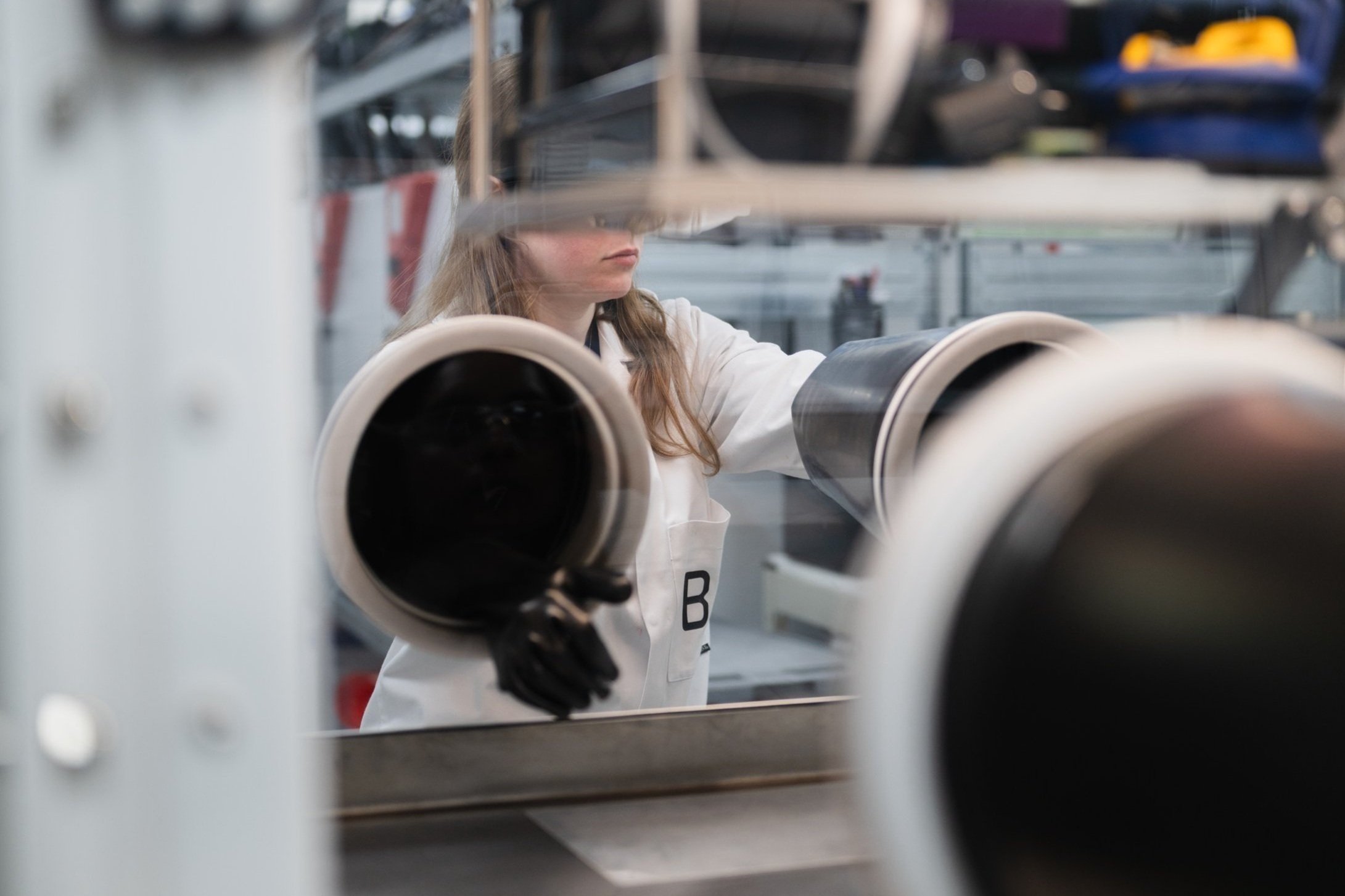 A woman inspecting or working on a large cylindrical object in an industrial or manufacturing setting, with shelves and tools visible in the background.