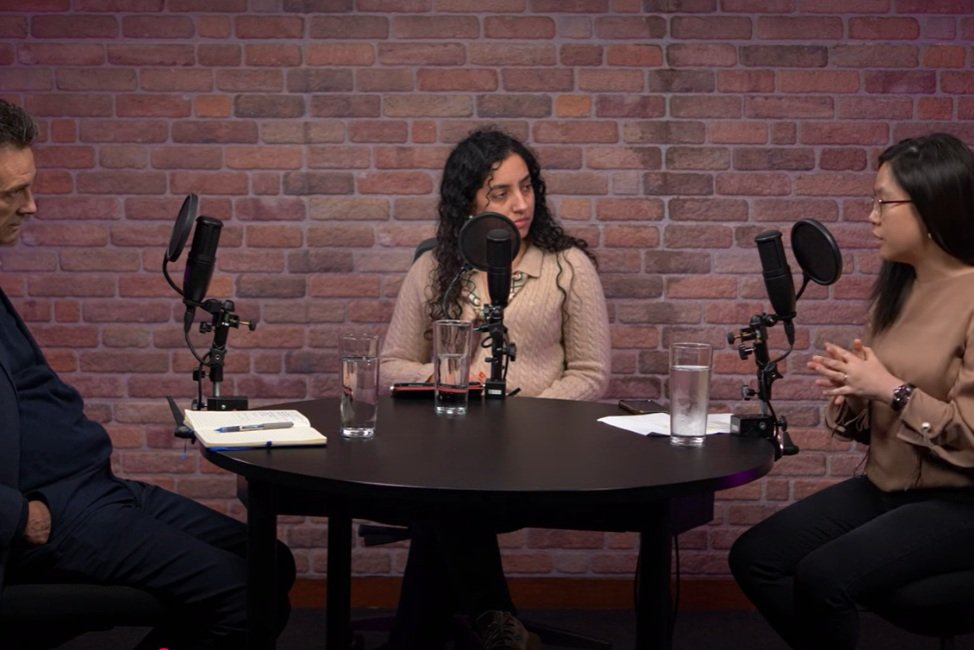 Three people sitting at a round table with microphones, glasses of water, and notebooks, engaged in a podcast recording against a brick wall backdrop.
