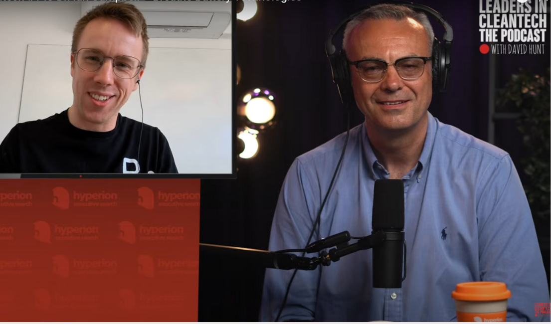 Two men participating in a podcast recording, one on a computer screen and one in a studio. The man on the screen has short hair, glasses, and is smiling. The man in the studio has short gray hair, glasses, and is wearing a light blue shirt, smiling at the camera. There is a microphone in front of him and a coffee cup on the desk. Background shows a studio setting with lighting and microphones.